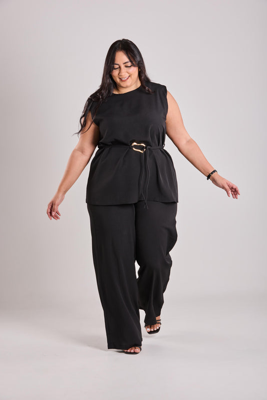 Woman wearing a black sleeveless top and pants with a belt on a light gray background